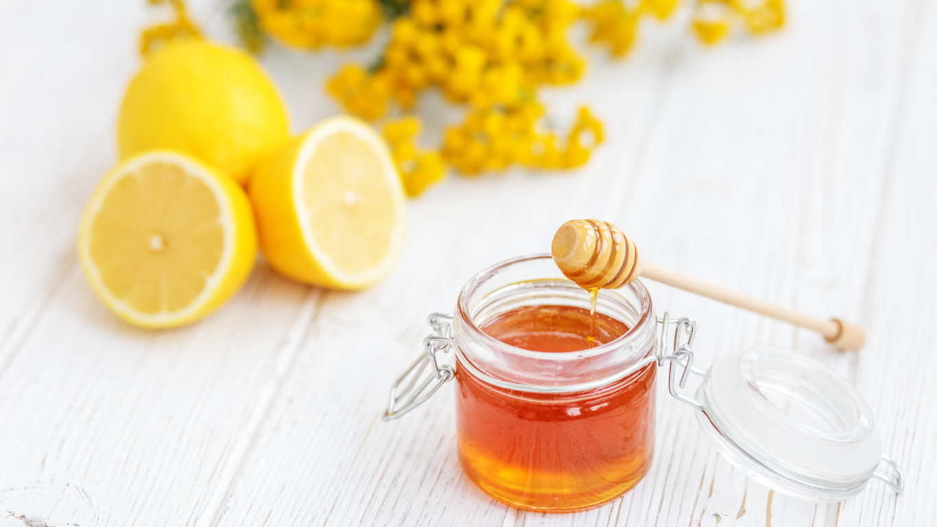 A jar of honey with a honey dipper, surrounded by flowers and herbs.